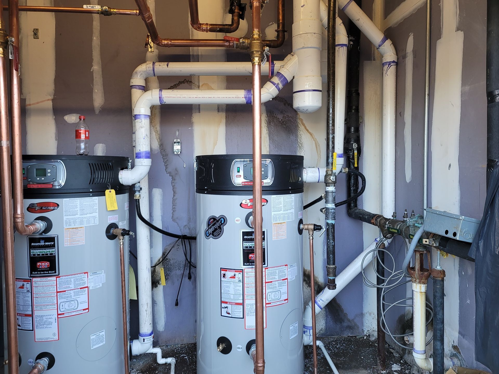 Two cylindrical water heaters with plumbing, copper pipes, and white PVC in a utility room.