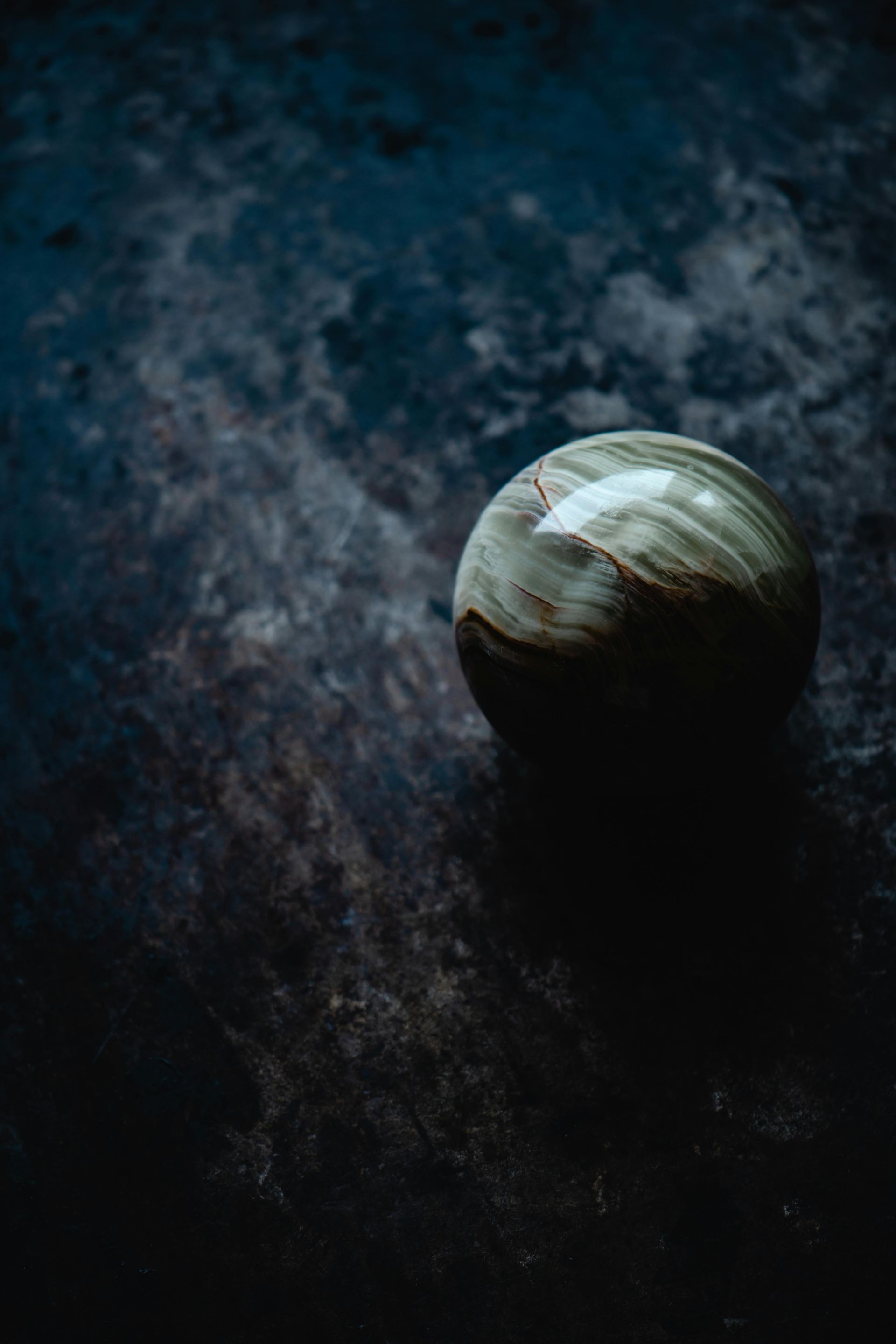 A smooth, spherical stone with swirling bands of green and brown rests on a dark, textured surface.