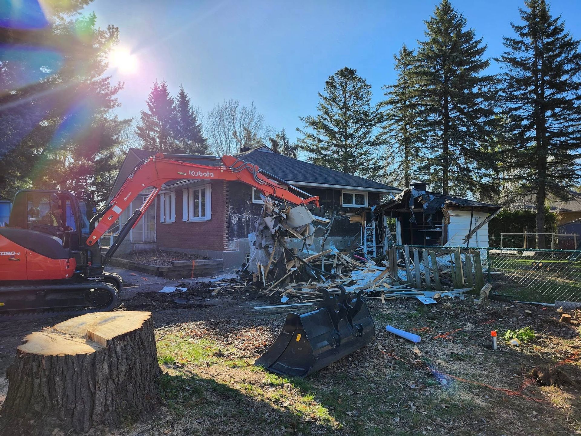 Une pelleteuse orange démolit l'extérieur d'une maison en briques au milieu d'une cour où se trouvent une grosse souche d'arbre et des pins.