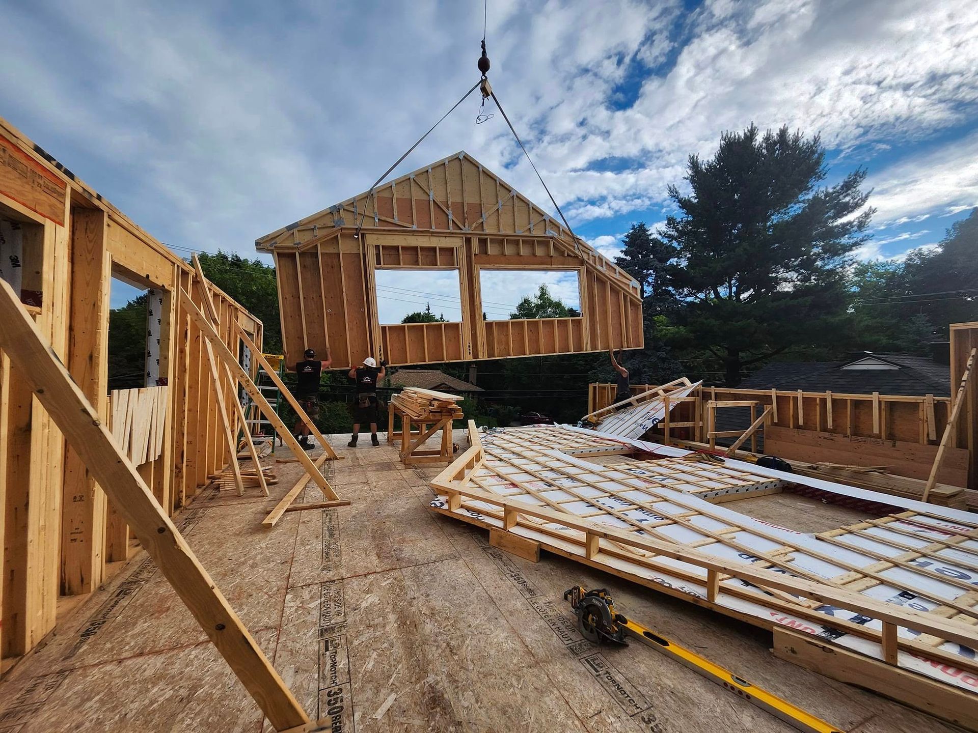 Sur un chantier de construction, une grue met en place une ossature murale en bois, tandis que des ouvriers se tiennent debout sur le sous-plancher en contreplaqué.