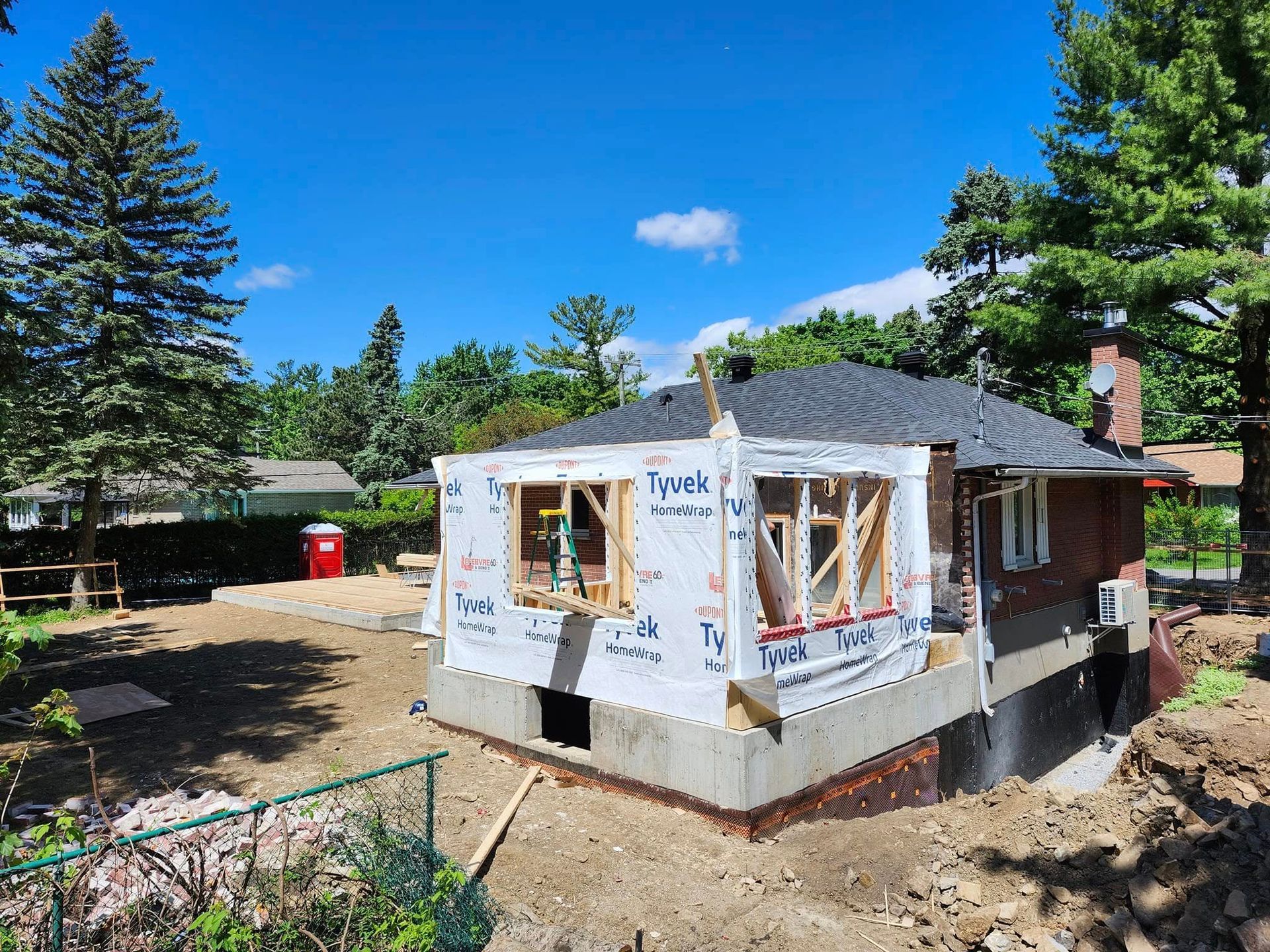 Une maison en construction avec des fondations en béton neuves et une ossature en bois recouverte de bâches Tyvek blanches.