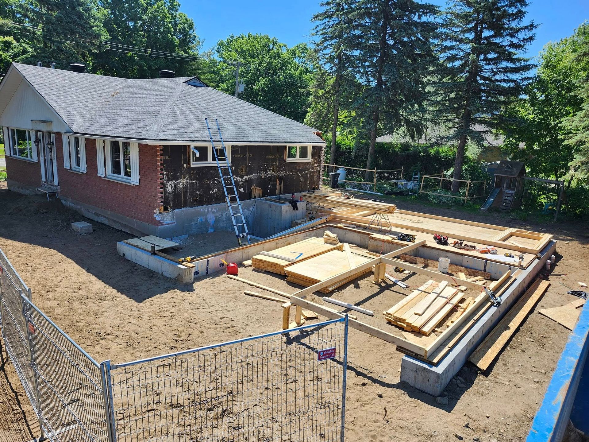 Chantier de construction montrant une maison en briques en cours d'agrandissement avec de nouvelles fondations en béton et une charpente en bois.