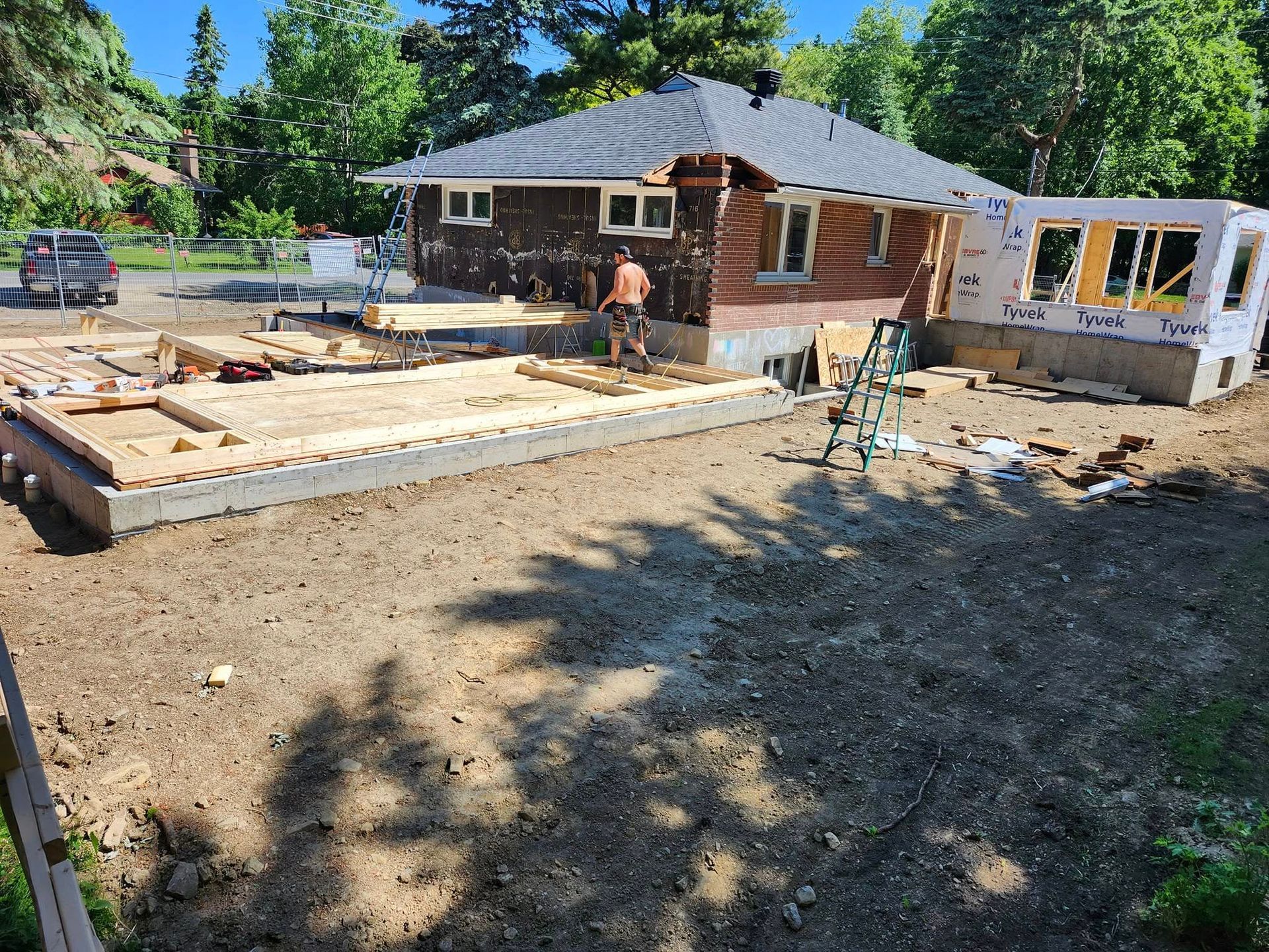 Un ouvrier du bâtiment construit une extension en bois sur la façade d'une maison, sous un ciel bleu dégagé.