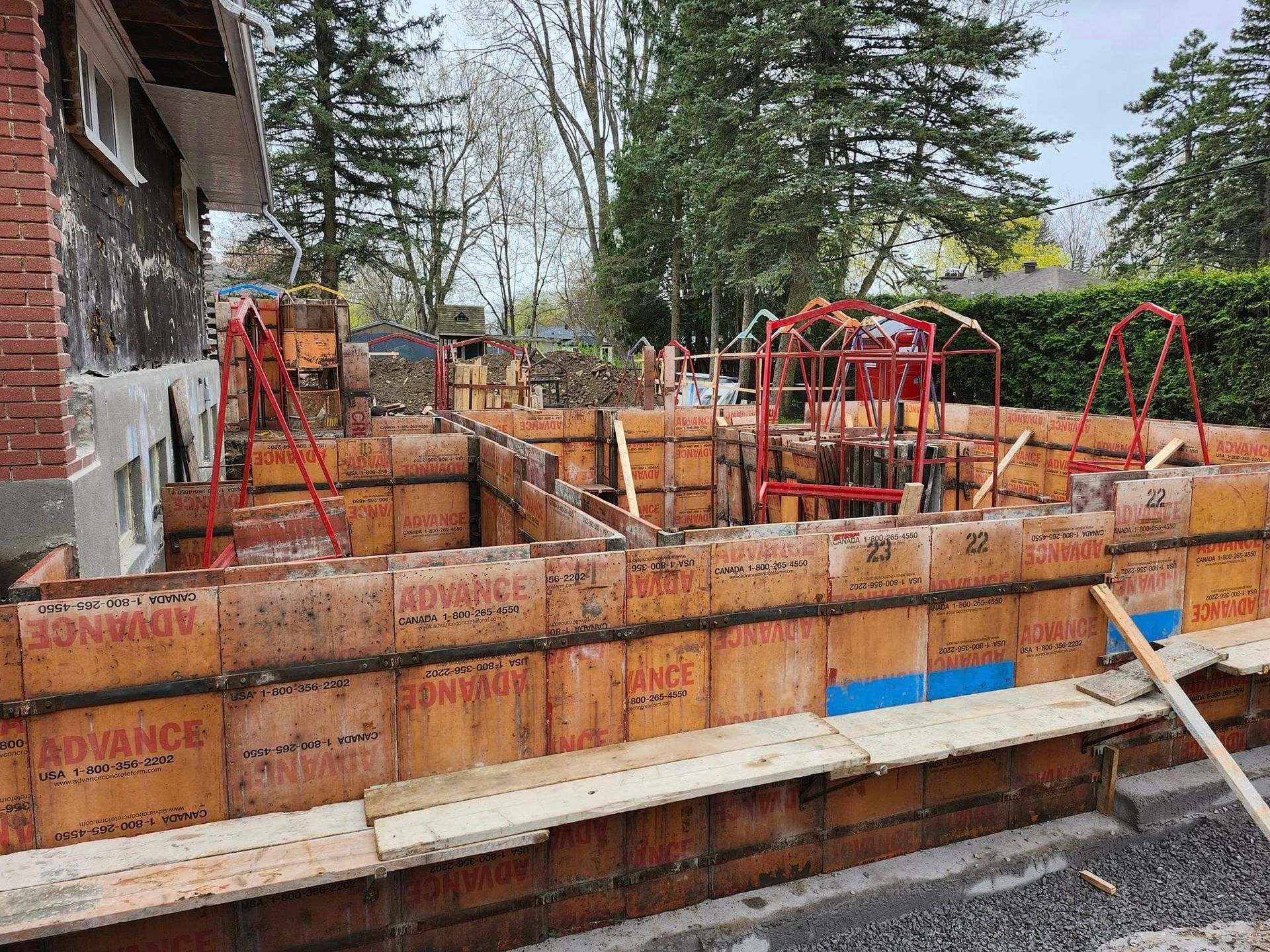 Chantier de construction montrant des coffrages modulaires en béton brun installés pour une fondation, avec des armatures en acier et un mur extérieur de maison.