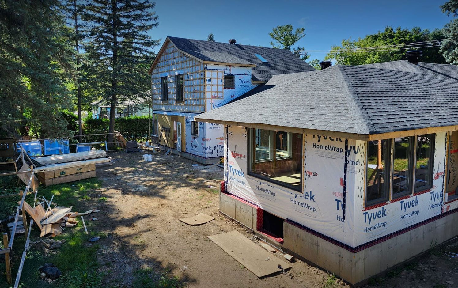 Un chantier de construction montre une extension de maison partiellement construite, avec un revêtement extérieur en Tyvek et un toit en bardeaux gris, par une journée ensoleillée.