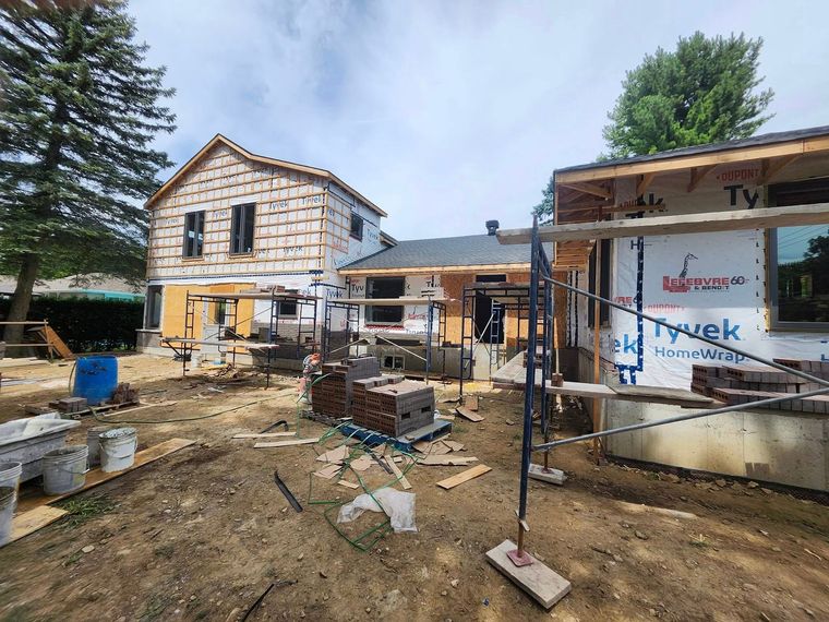 Un chantier de construction montrant deux parties de maison inachevées en cours de rénovation, avec une charpente en bois apparente et un bardage blanc.