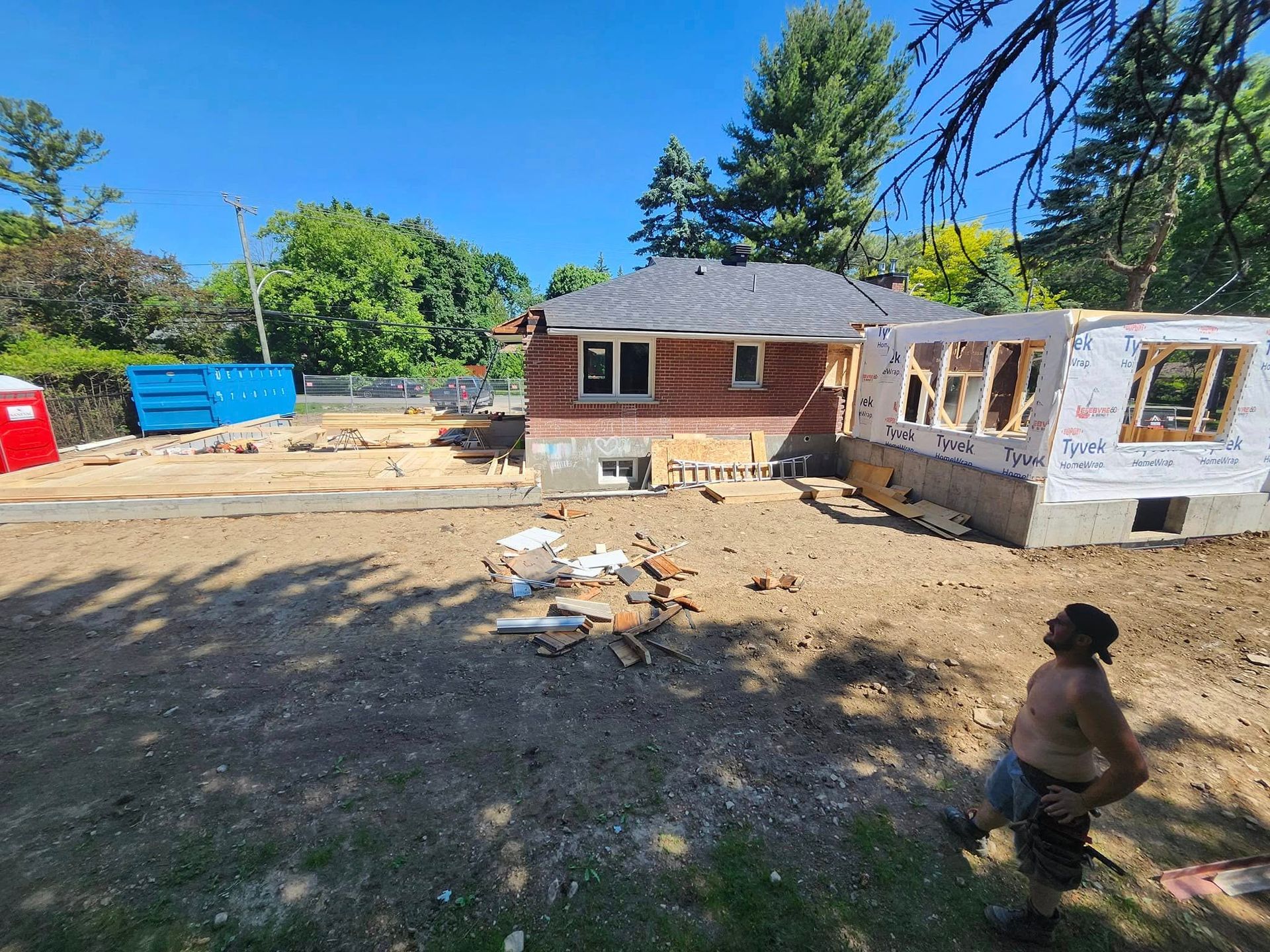 Une personne se tient dans une cour en terre battue et regarde une maison en rénovation, avec une nouvelle charpente et une benne à ordures bleue à proximité.