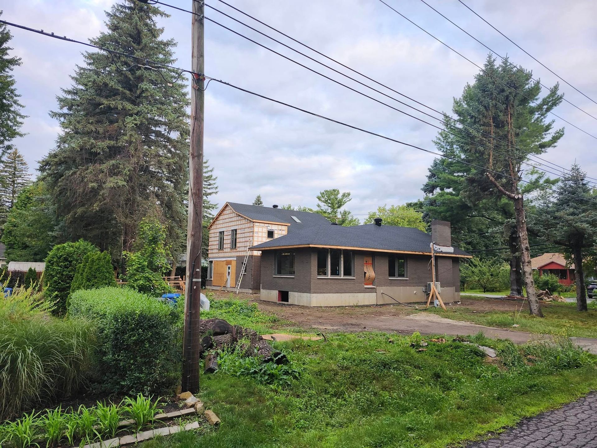 Une maison en construction, avec un revêtement extérieur sombre, un toit en bardeaux et des murs inachevés, dans un jardin boisé et verdoyant.