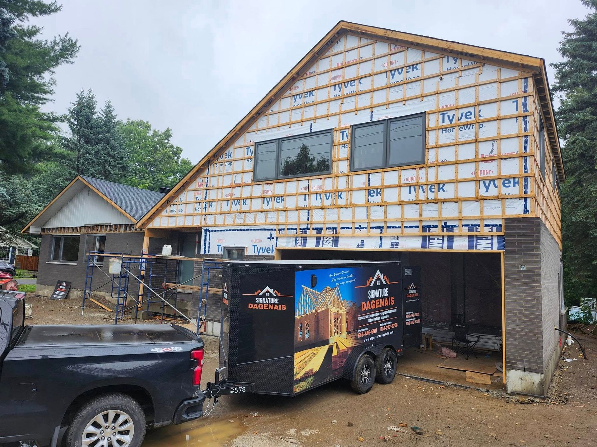 Un camion noir tractant une remorque de chantier de marque est stationné devant une maison en rénovation avec un revêtement Tyvek.
