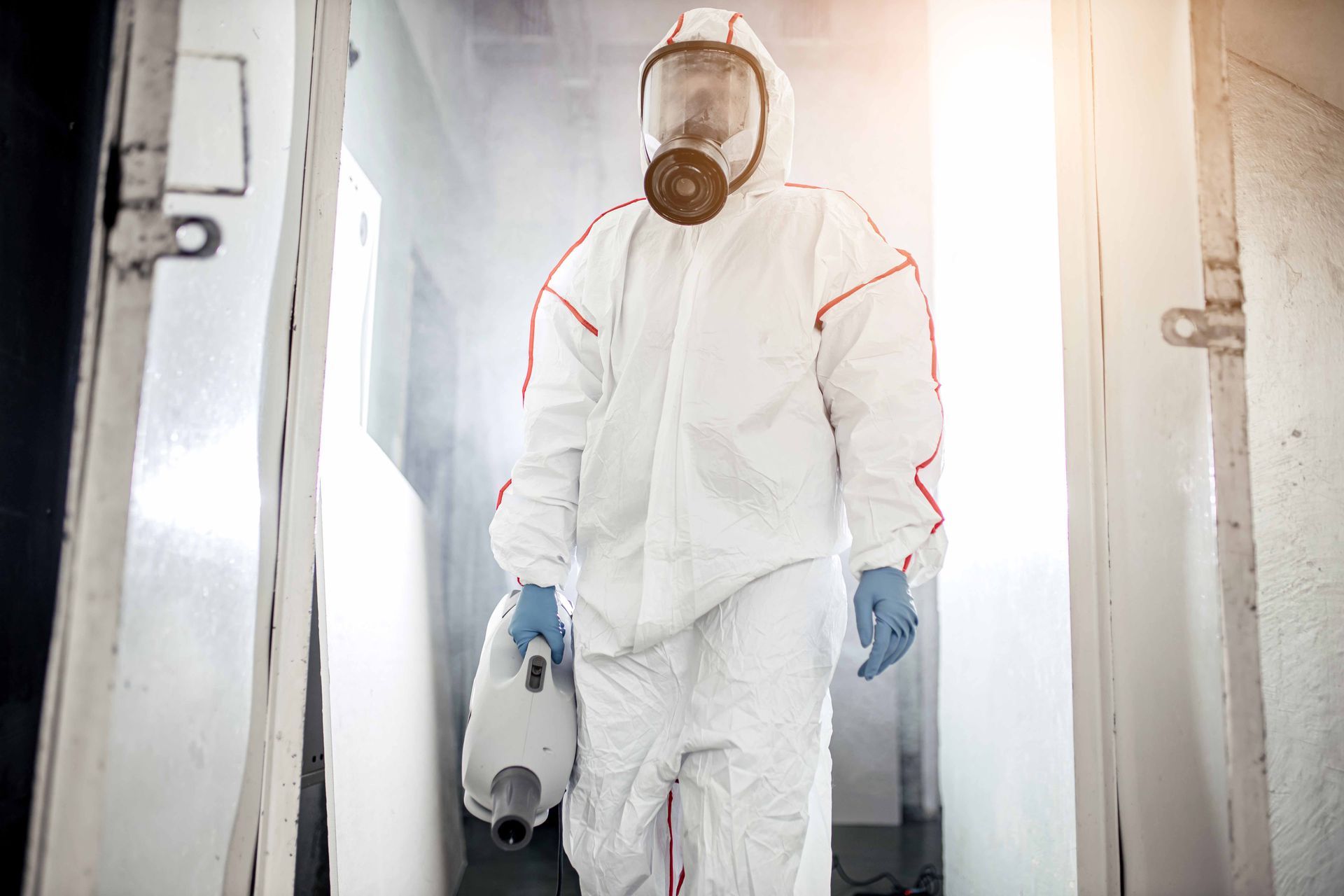 Une personne en combinaison de protection et masque respiratoire sort d'une porte, tenant un nébuliseur.