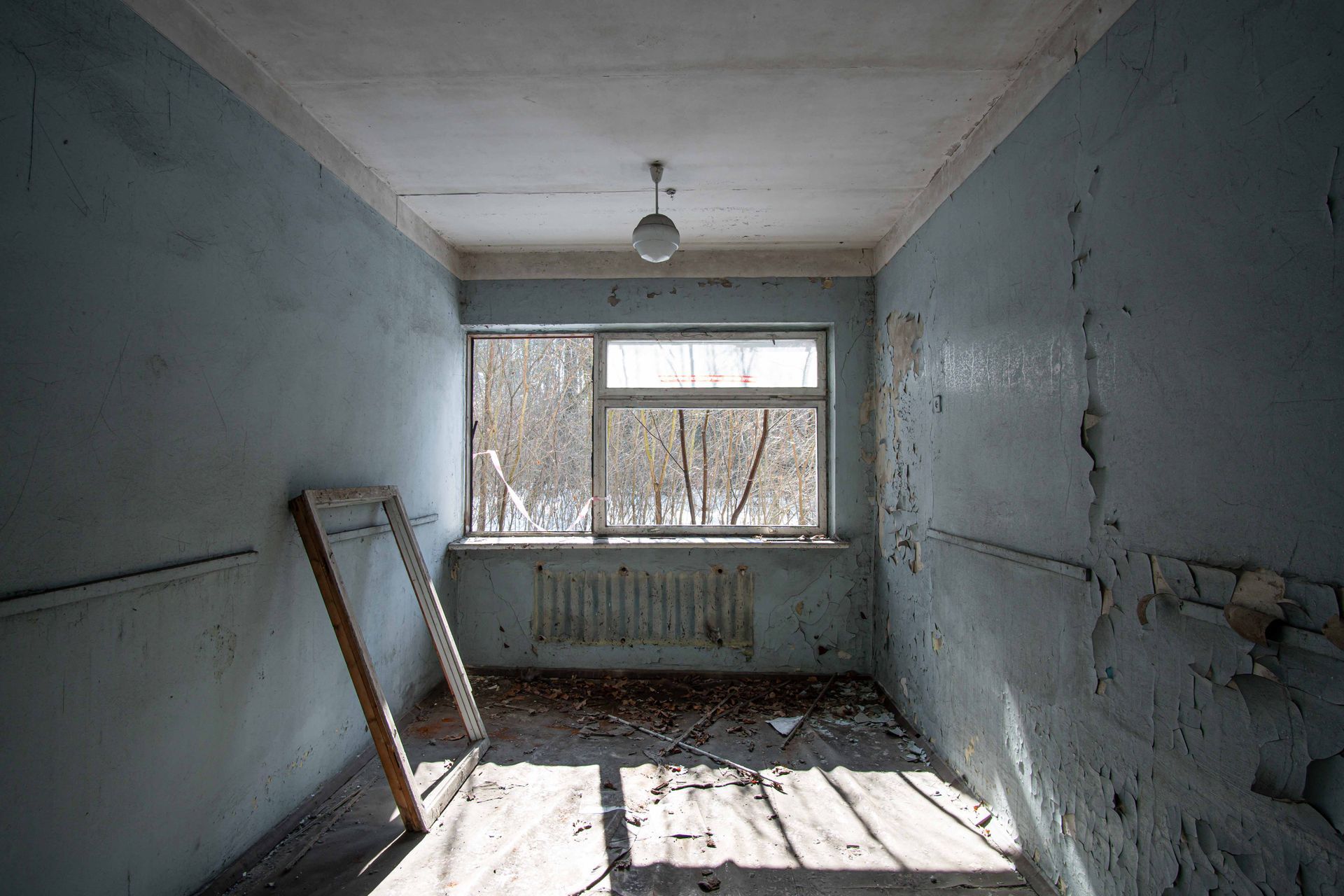 Pièce abandonnée et délabrée, aux murs bleus écaillés, avec une fenêtre et des débris au sol.