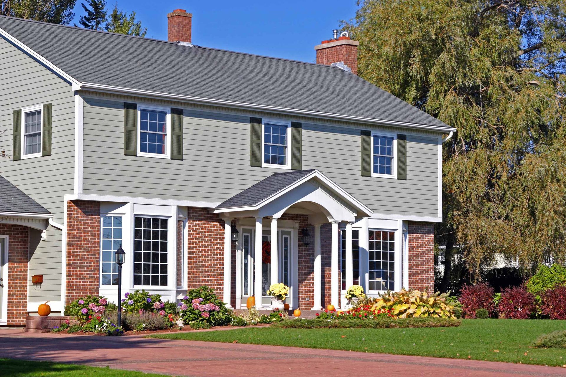 Two-story house with green siding, brick accents, white trim, and a gray roof, in a sunny setting with landscaping.