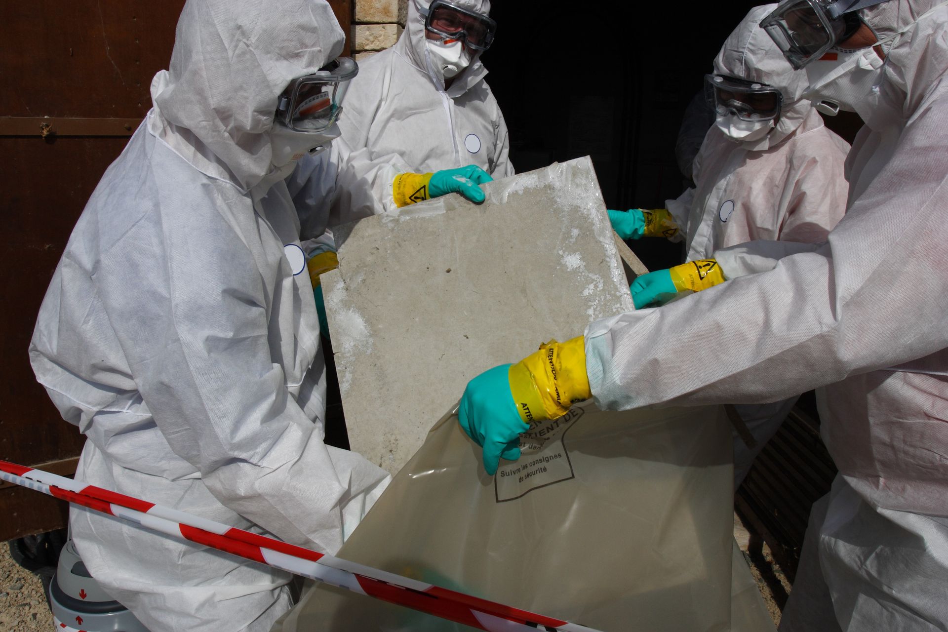 People in protective suits placing a cement slab into a bag, possibly asbestos removal.