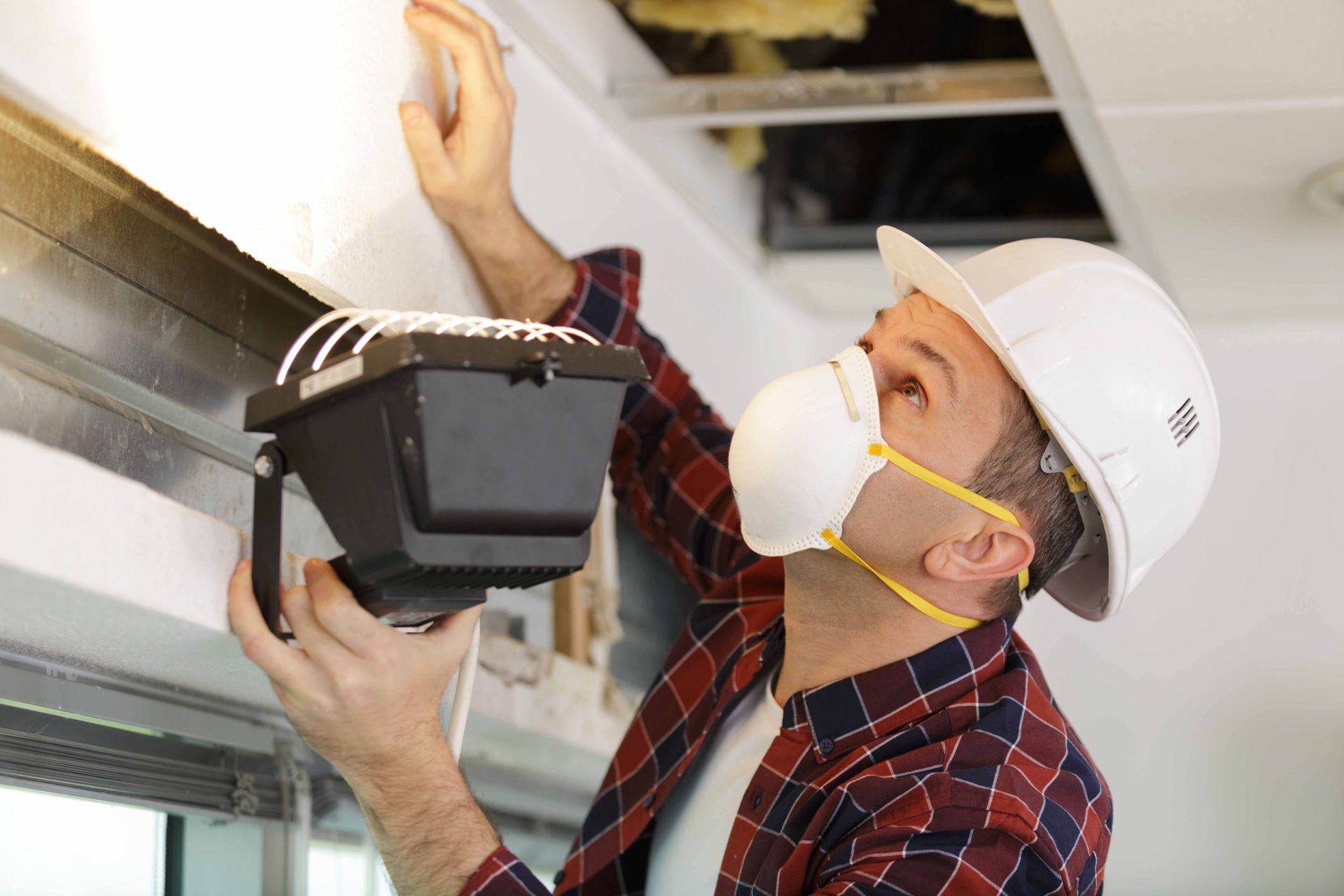 Un homme portant un casque et un masque inspecte un plafond, tenant une lampe torche, regardant vers le haut.