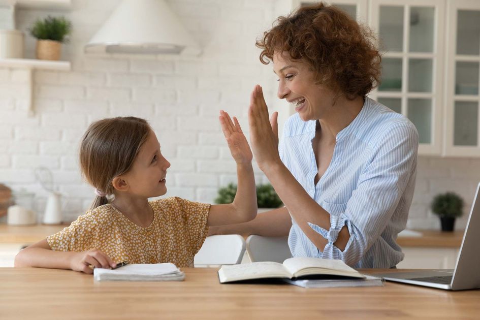 Adult and child high-fiving at a table with open books in a bright home kitchen