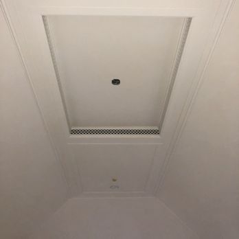 A white recessed ceiling panel with molding, a small dark light fixture, and a rectangular metal ventilation grate.