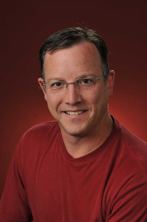 Smiling man in glasses and a red shirt against a dark red background