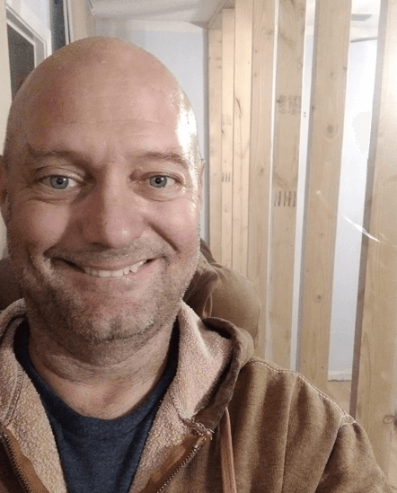A bald man with a beard is smiling in front of a wooden wall.