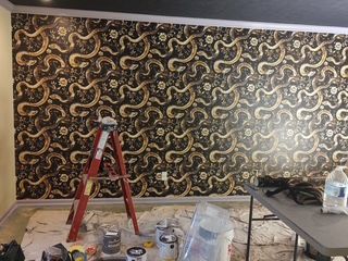 Room with black-and-gold patterned wallpaper, red ladder, and painting supplies on the floor.