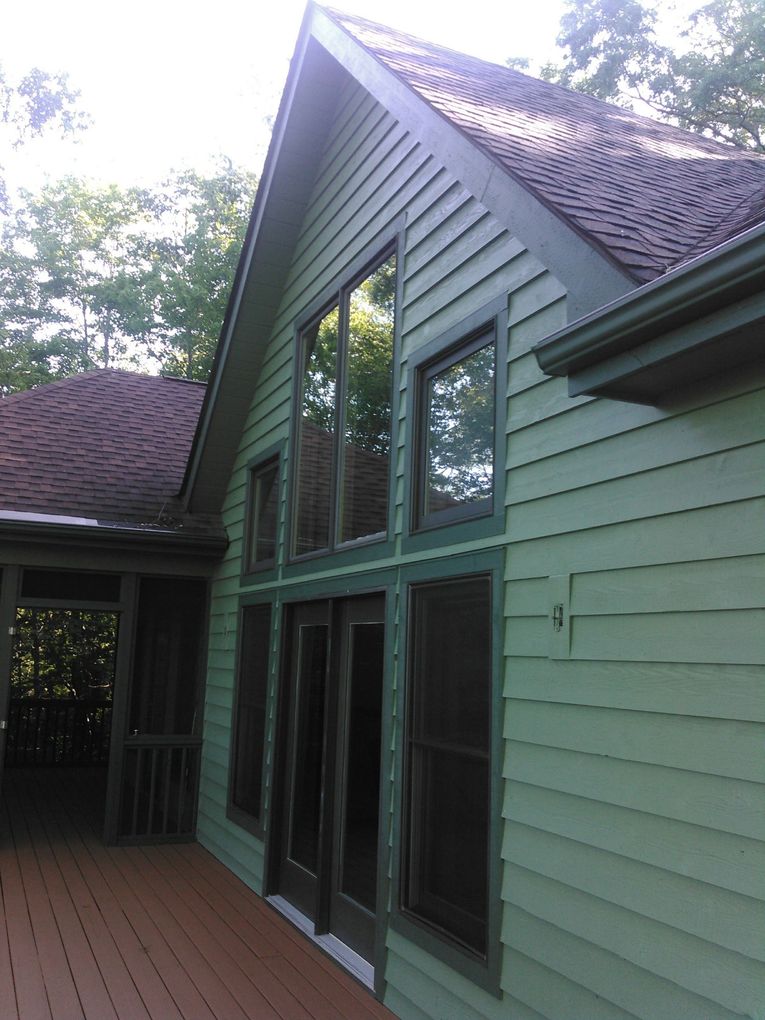 Green house exterior with large windows, red shingle roof, and wooden deck under trees