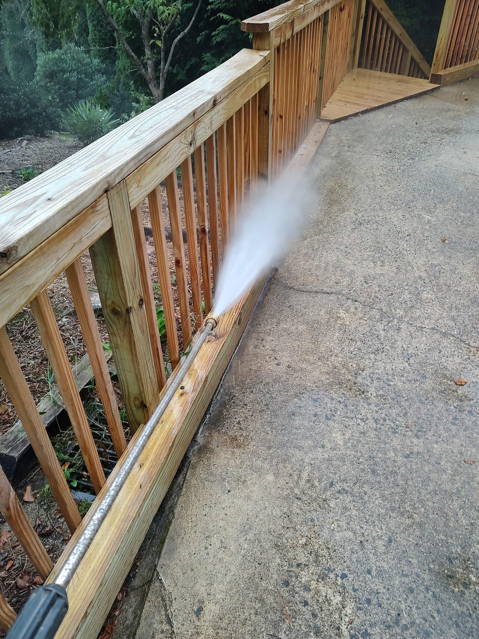 Person pressure-washing a wooden deck railing beside a gravel path, with water spraying onto the boards