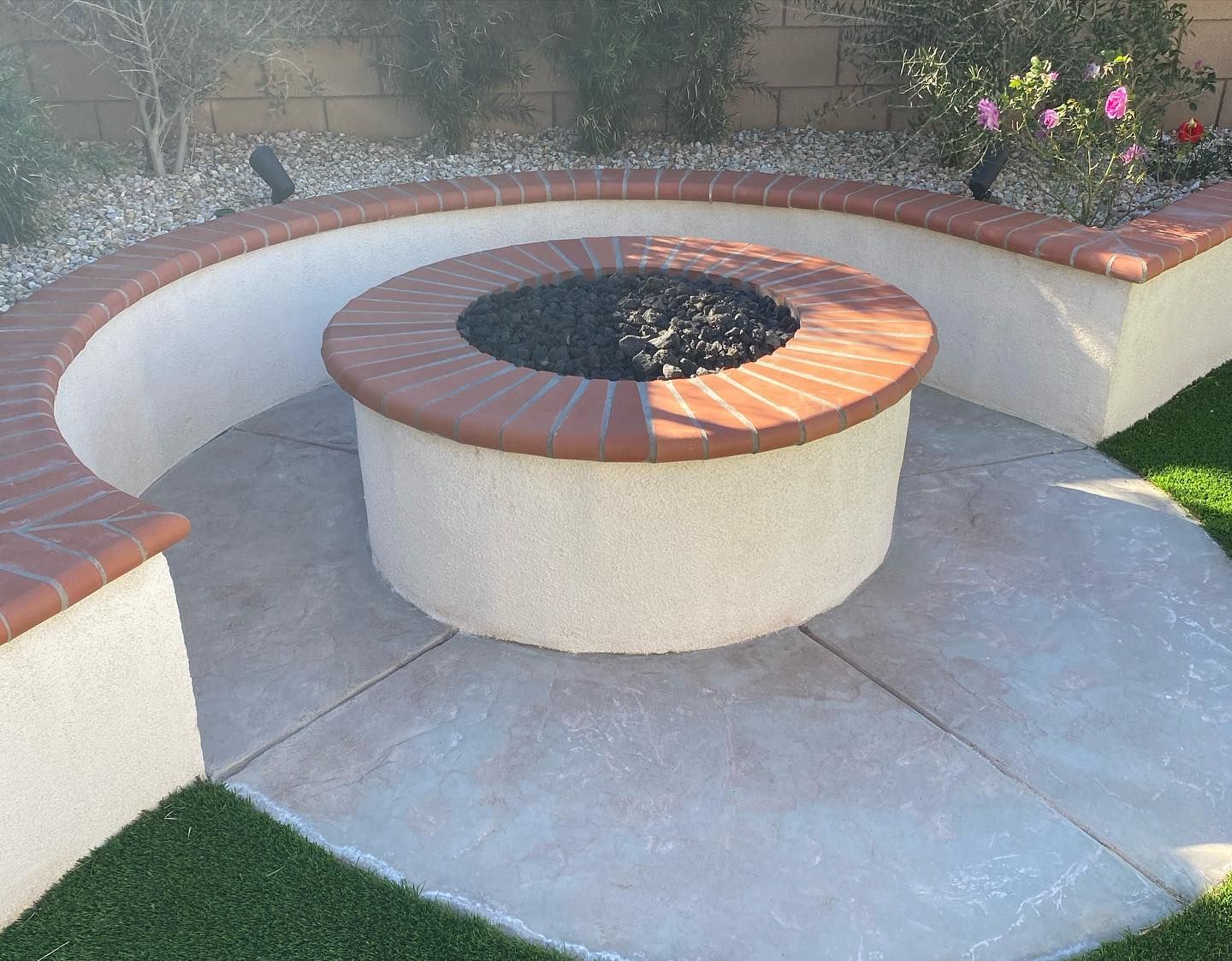 Curved outdoor bench around a circular fire pit in a landscaped patio with white walls and flowers