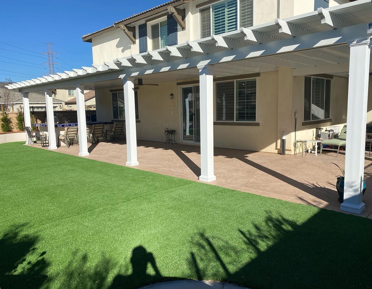 Patio with white pergola columns beside a two-story house and green lawn in bright sunlight