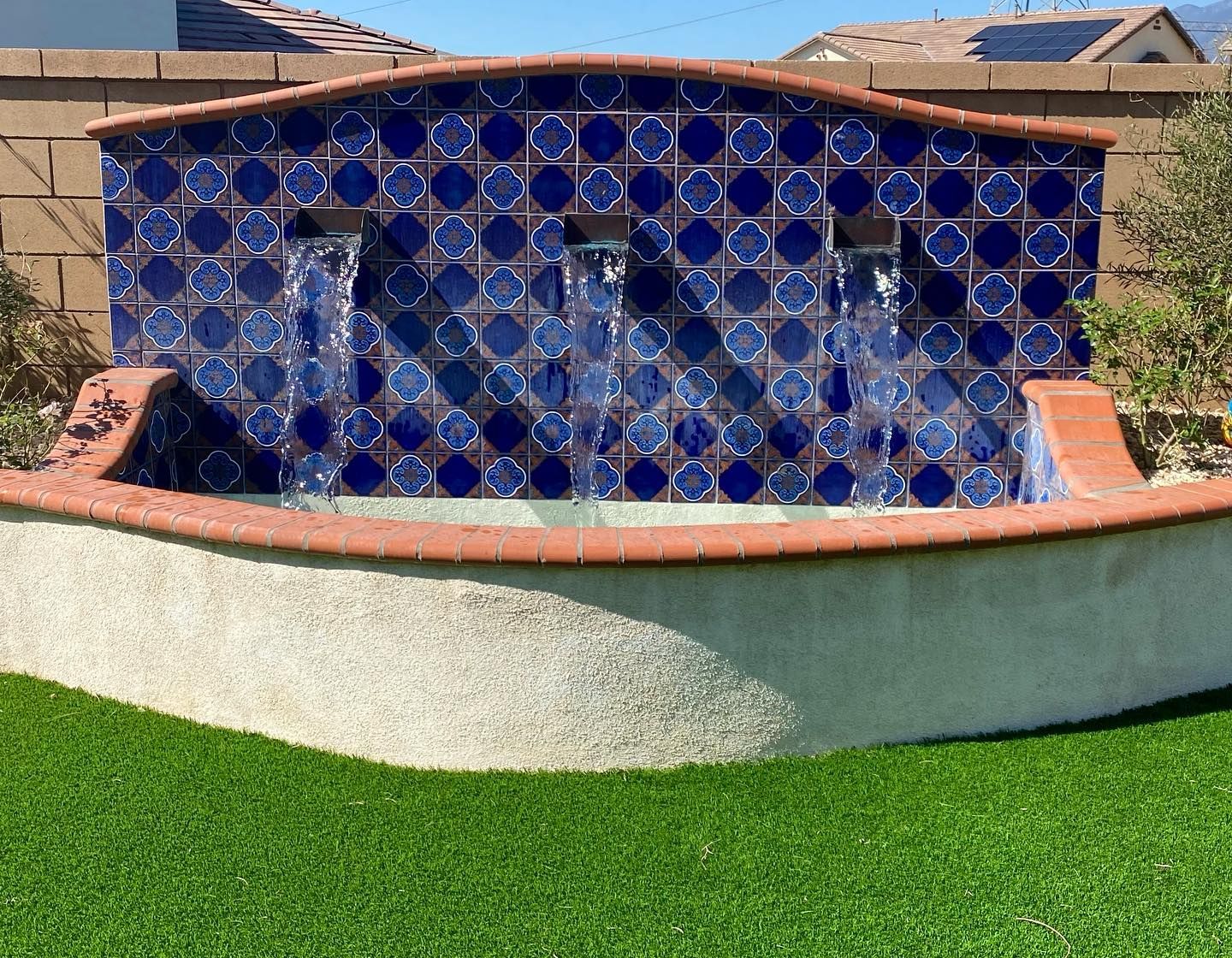 Blue tiled wall fountain with water cascading over a curved stucco base in a landscaped yard