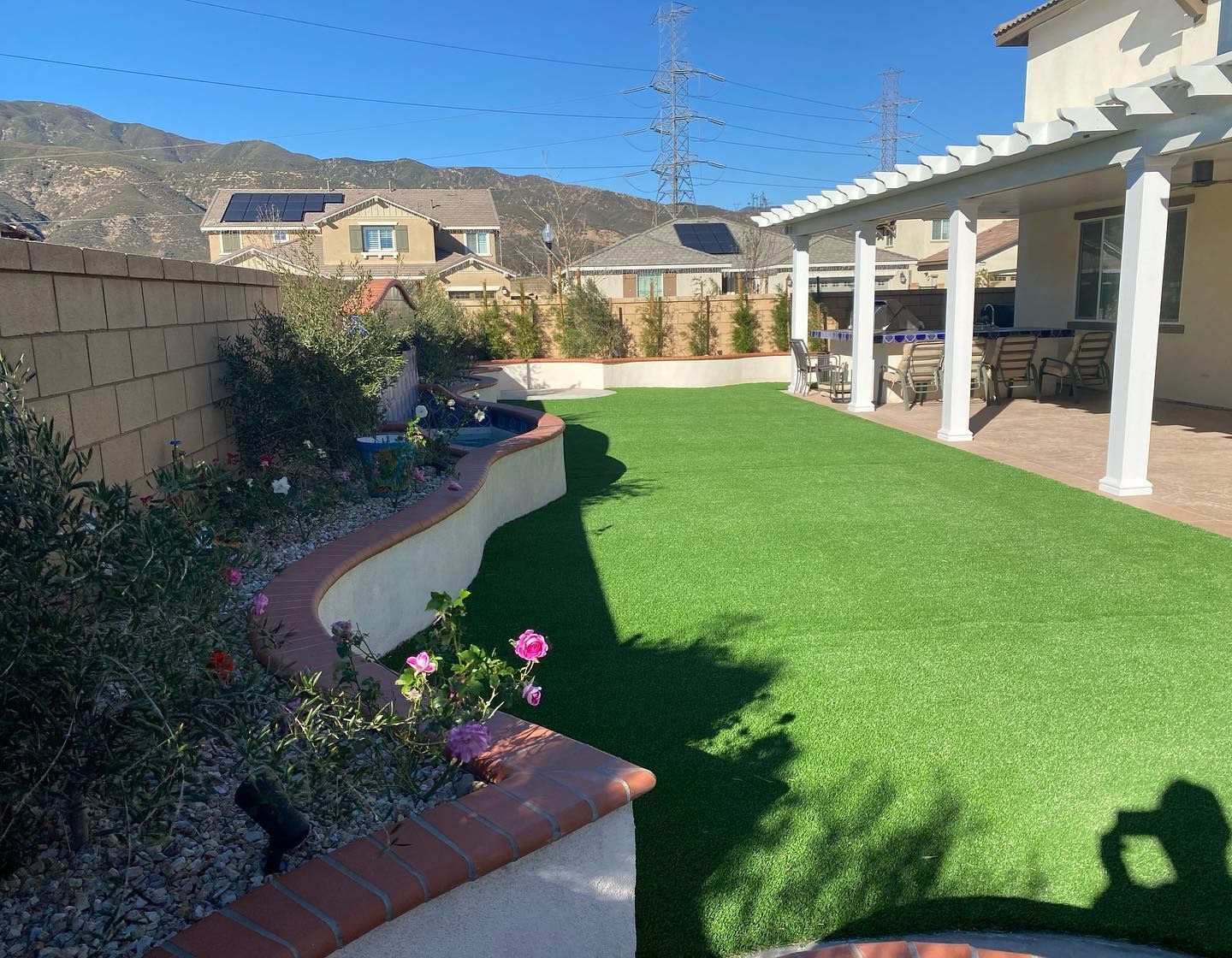 Sunny backyard with green lawn, flower bed, and covered patio beside desert hills