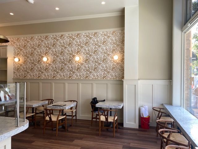 Quiet café interior with floral wallpaper, wall sconces, wooden tables, and chairs near a window.