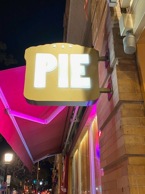 Lit “PIE” sign on a brick storefront at night, with a pink awning and streetlights.