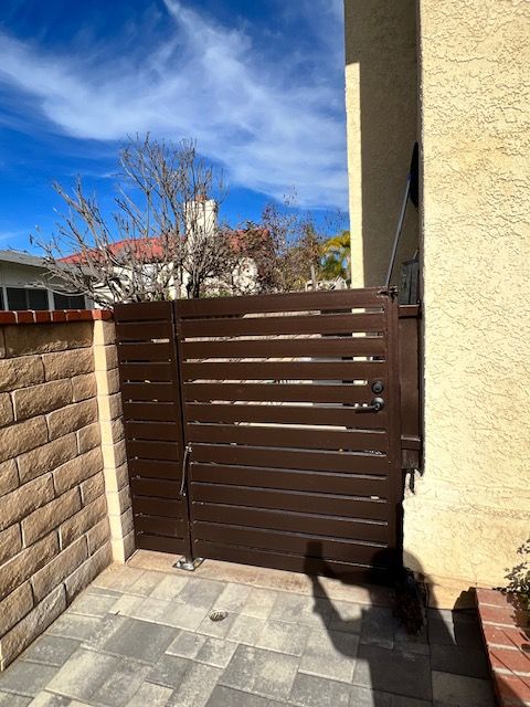 Brown slatted gate in a small sunlit patio beside a beige stucco wall and brick planter.