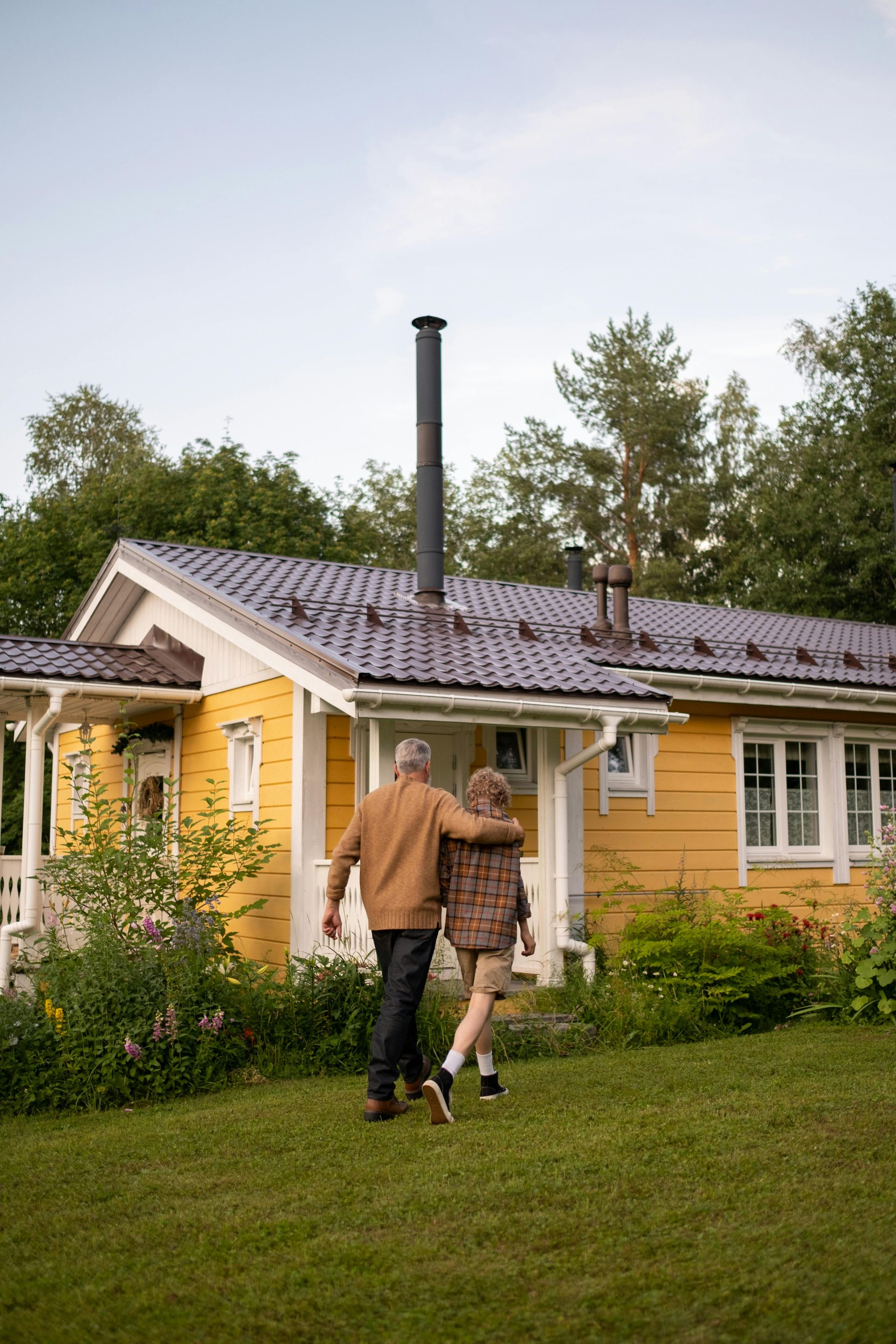 Couple walking toward a yellow cottage with a smoking chimney in a grassy yard