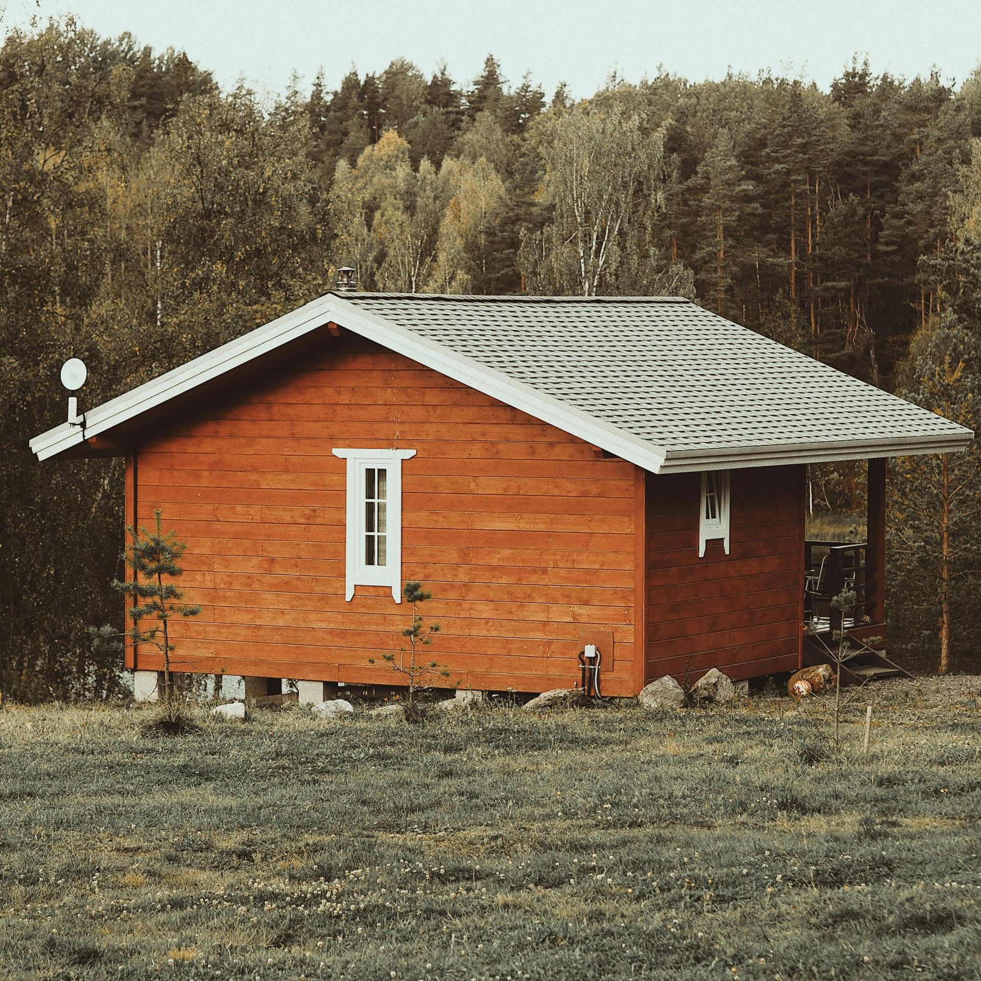 Small red wooden cabin with a gray roof in a grassy field, backed by dense forest.