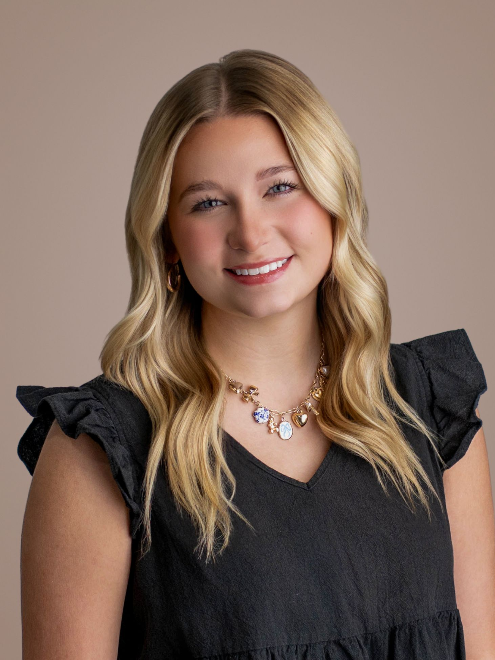 Smiling person with blonde hair wearing a dark ruffled top and necklace against a plain background