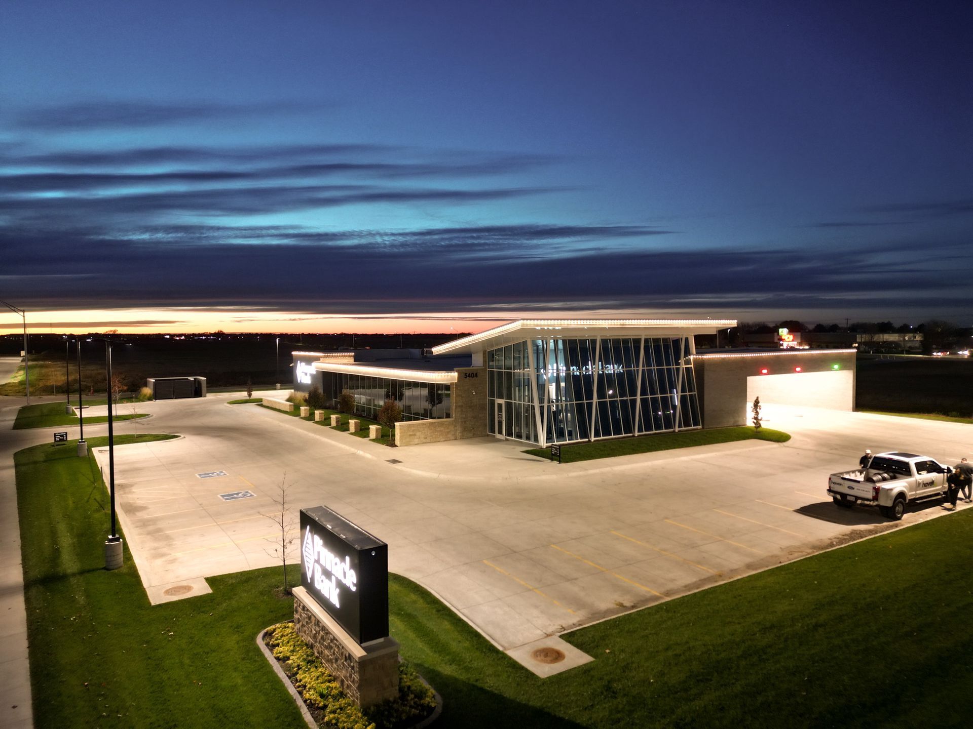 Modern glass-fronted building at dusk with parking lot and illuminated sign under a dark blue sky