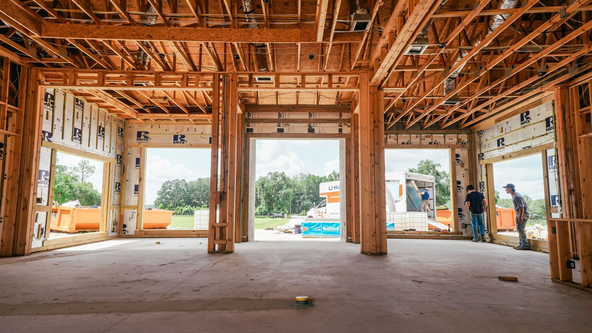 Unfinished house interior with exposed wooden framing and workers near open wall openings.