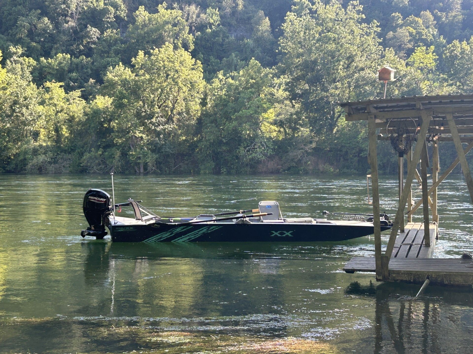 Small fishing boat on calm river beside a dock, with wooded shoreline in the background