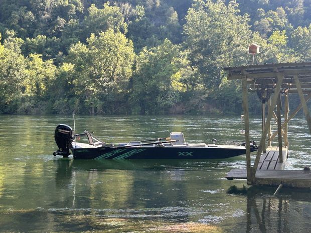 Small fishing boat on calm river beside a dock, with wooded shoreline in the background