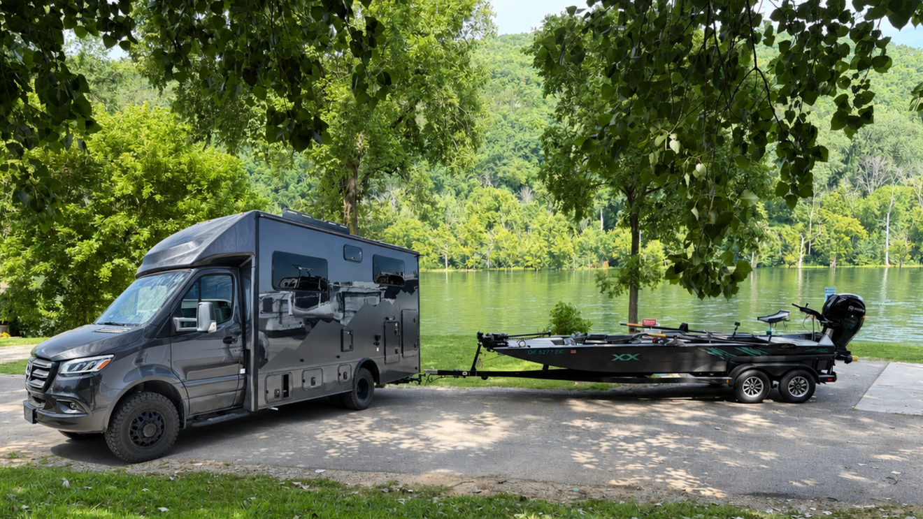 Gray camper van towing a boat trailer beside a calm lakeside under trees