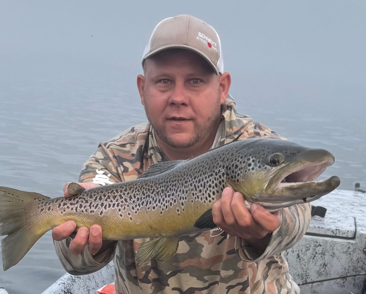 Angler holding a large spotted fish on a foggy lake in camouflage gear and cap