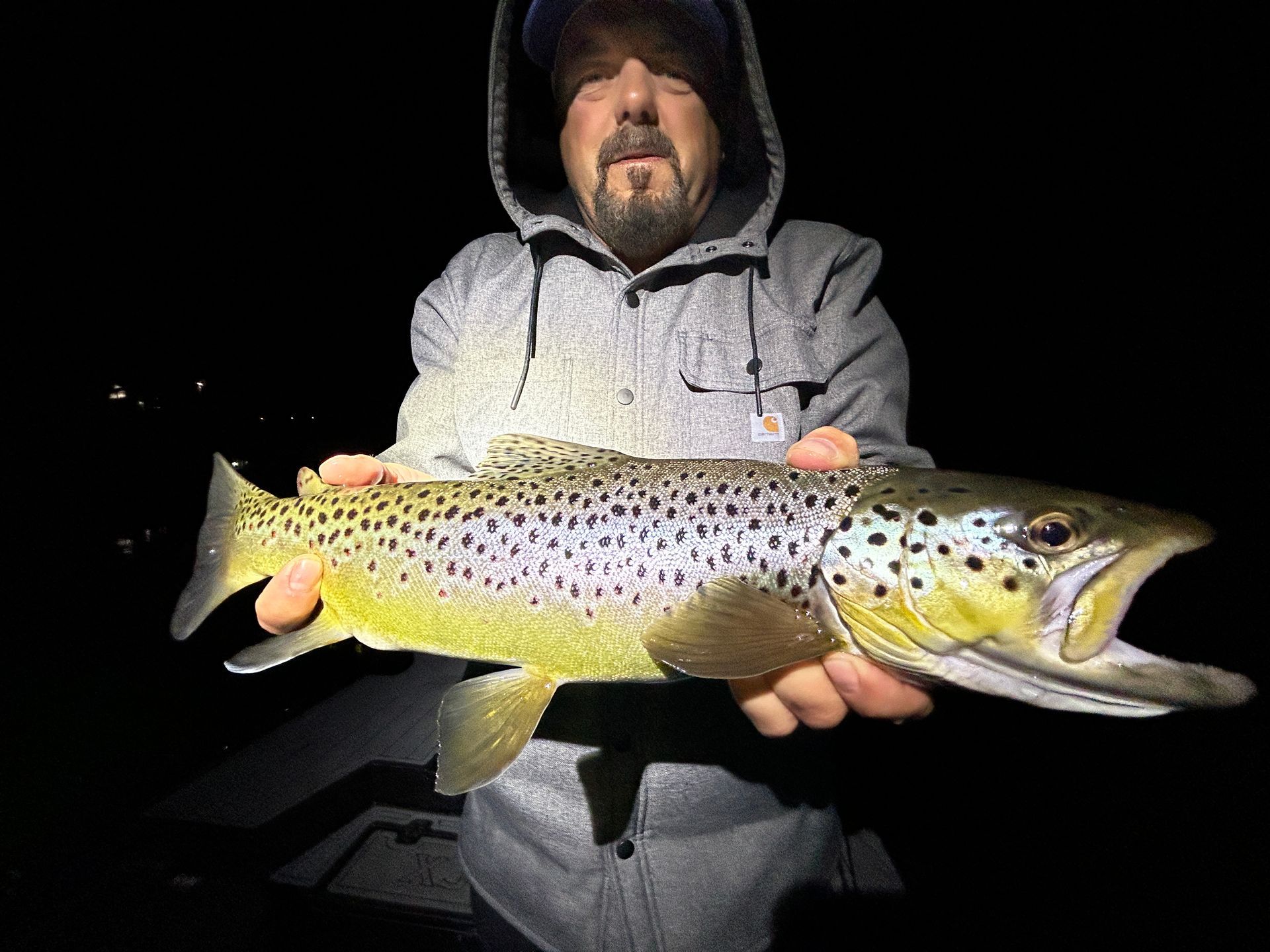 Man holding a large spotted fish at night, wearing a hooded jacket