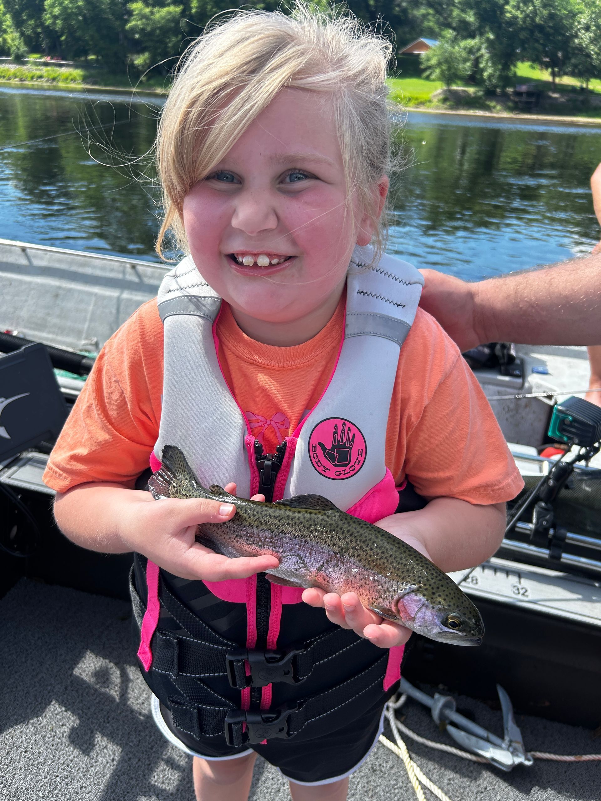 Smiling child holding a fish on a boat by a lake, wearing a life jacket.