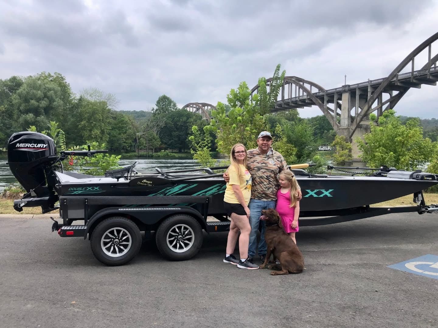People posing beside a black bass boat with a dog, on a road near a bridge and water.