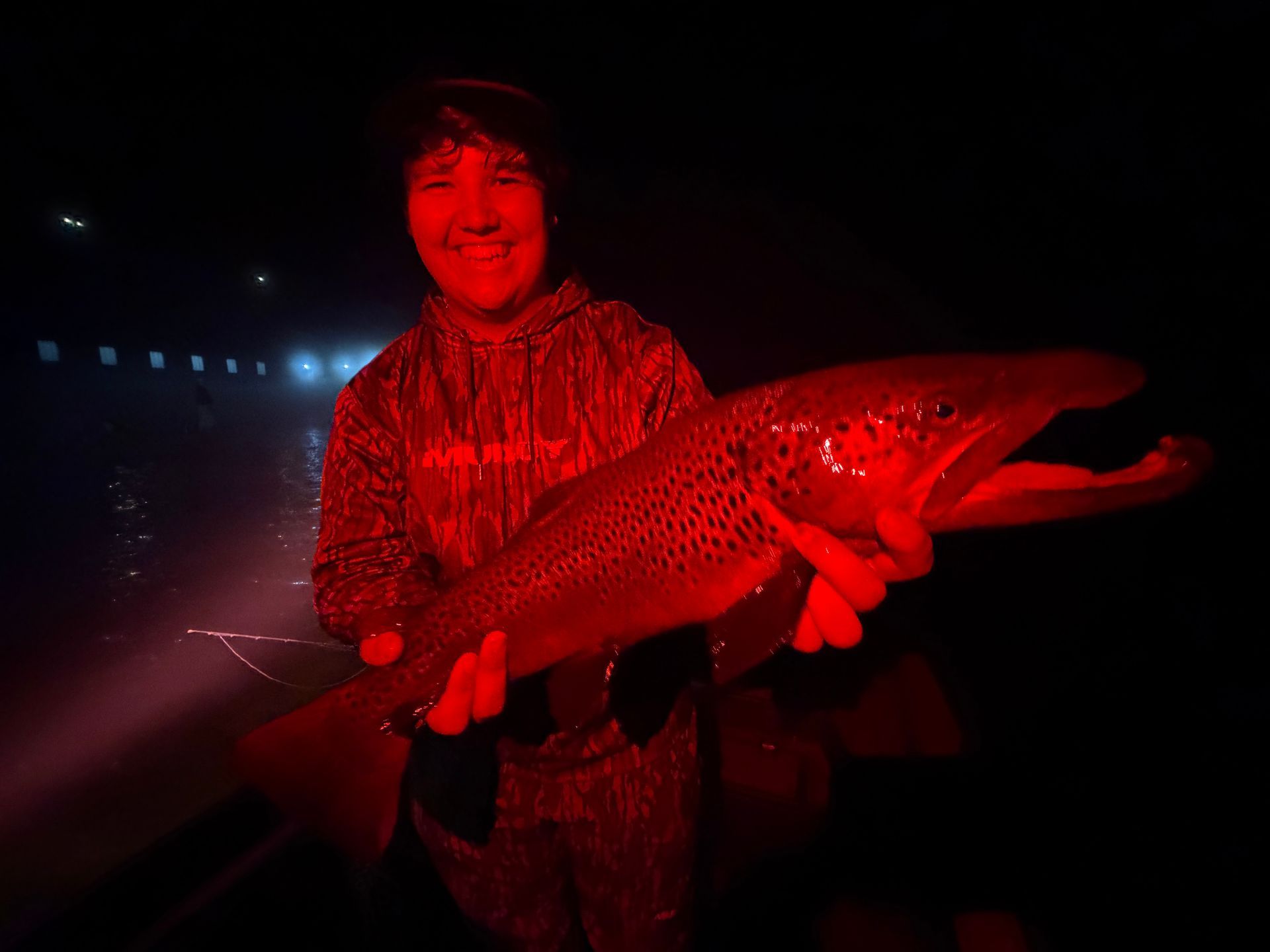 Person holding a large fish at night, lit by a bright red light near the water