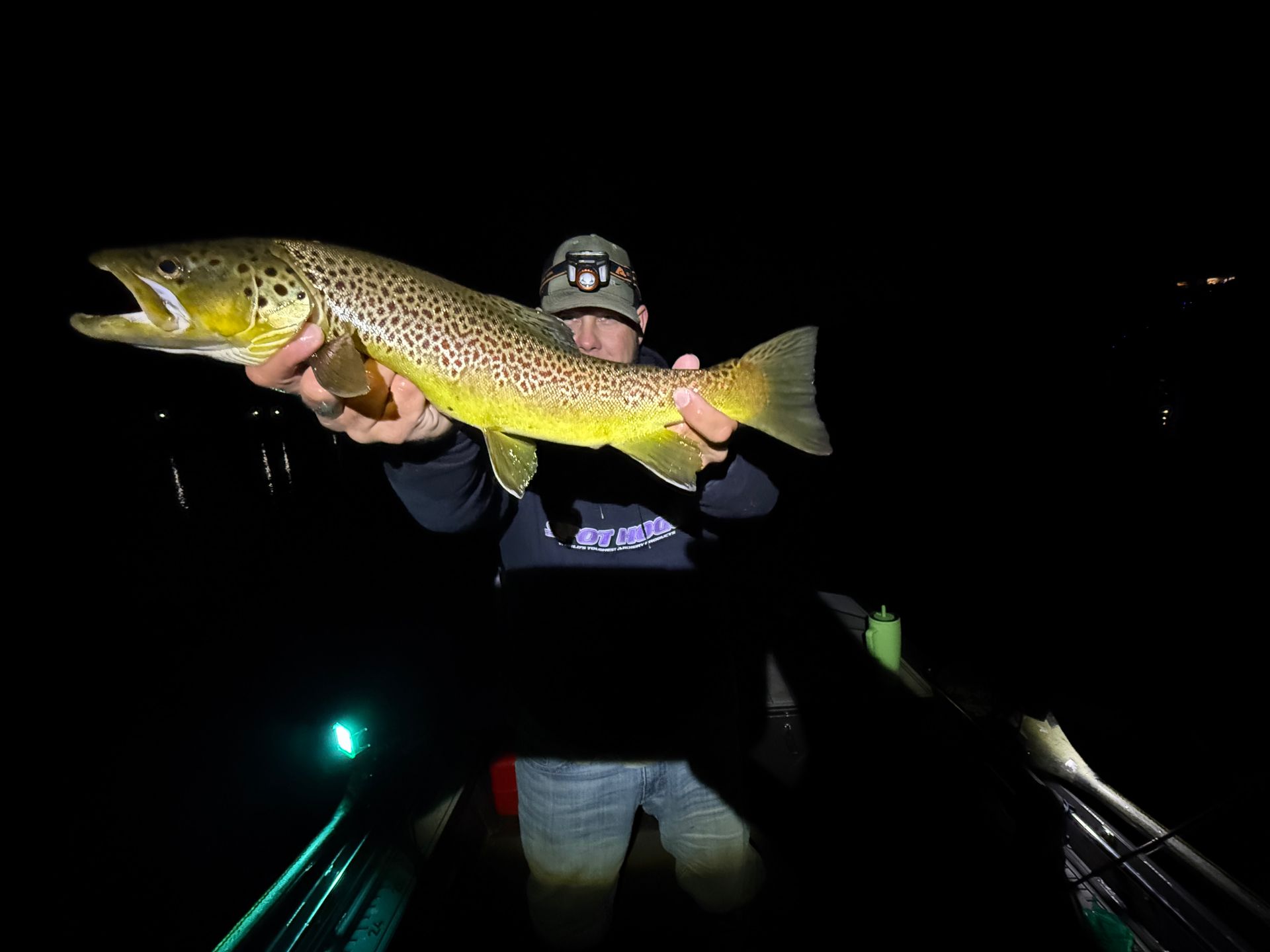 Angler holding a large spotted fish at night on a boat, with dark water and a headlamp glow