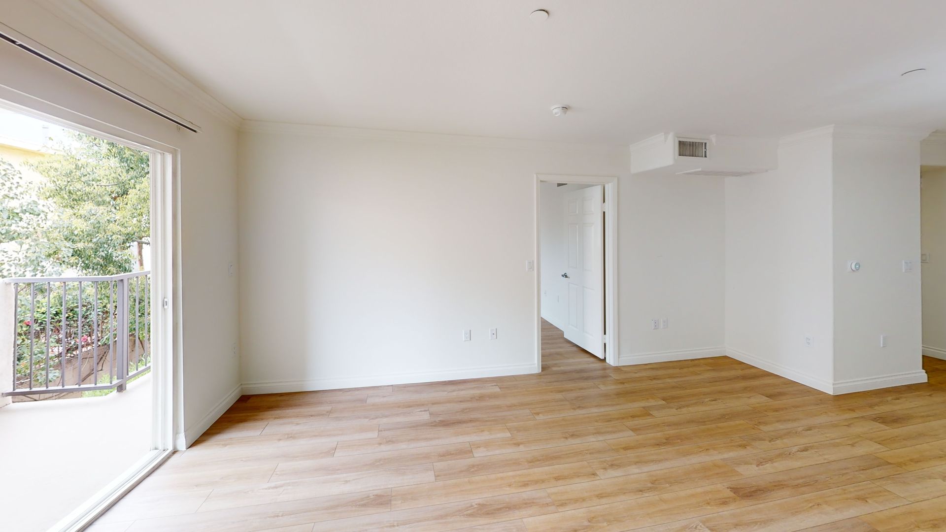 Empty room with light wood floors, white walls, and a balcony visible through a sliding glass door.