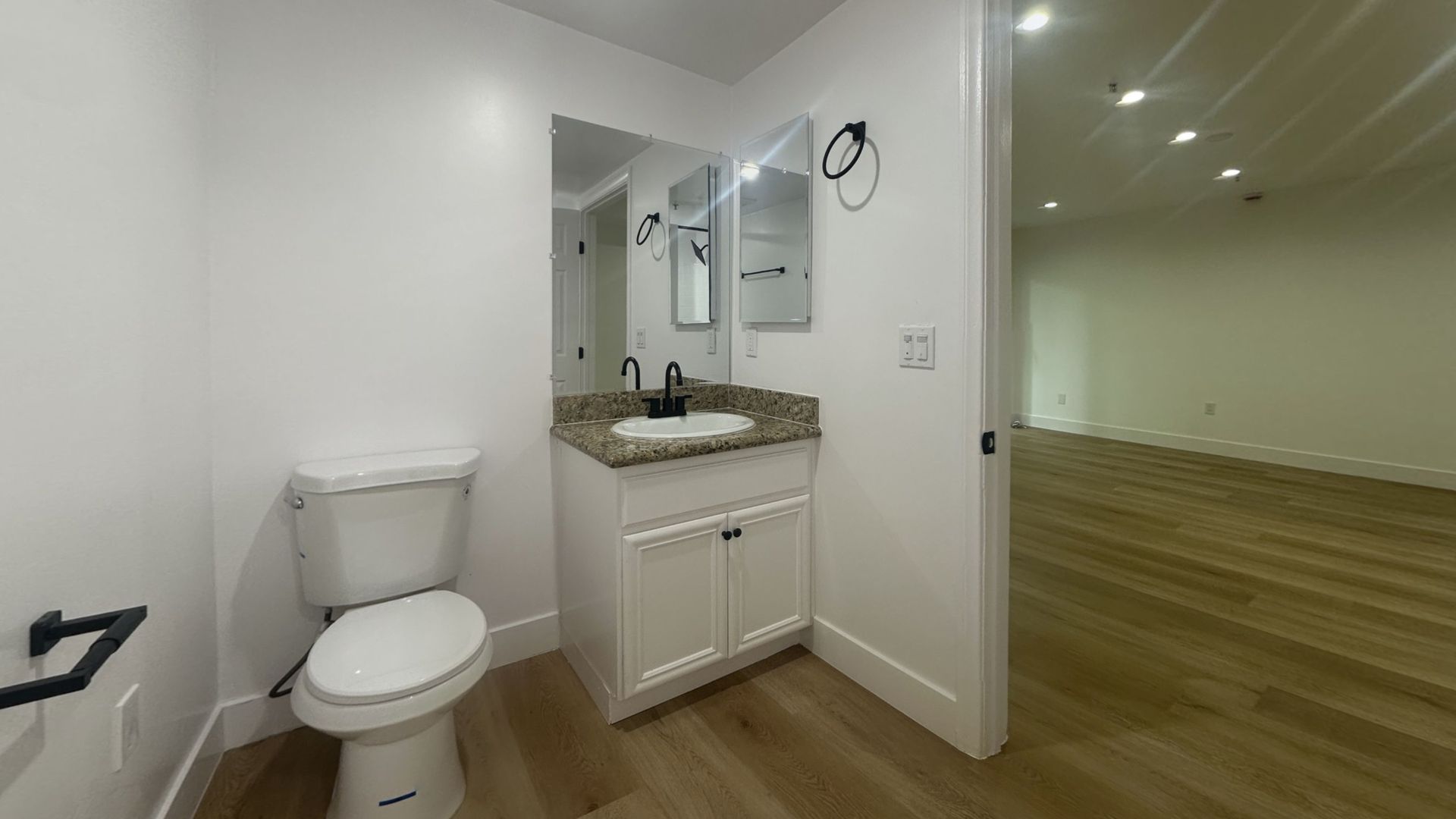 Bathroom with white toilet, vanity, and open doorway to a room with hardwood floors.