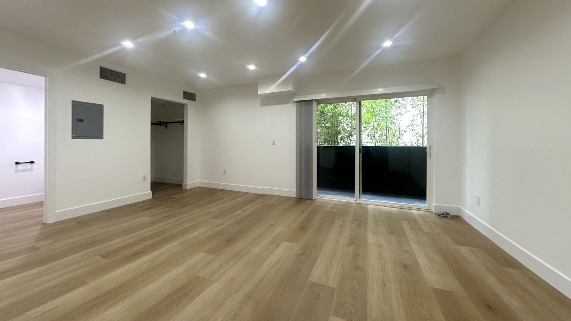 Empty, brightly lit room with hardwood floors, recessed lighting, and a sliding glass door leading to a balcony.