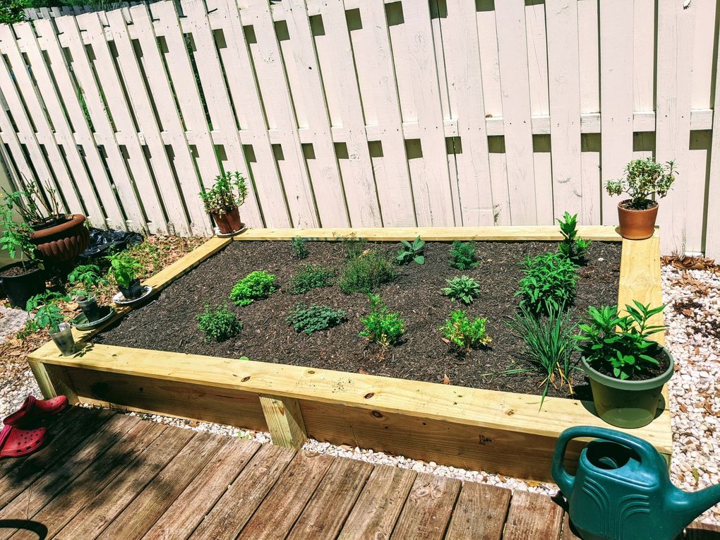 Wooden raised garden bed with green plants, on a wooden deck, near a white fence.